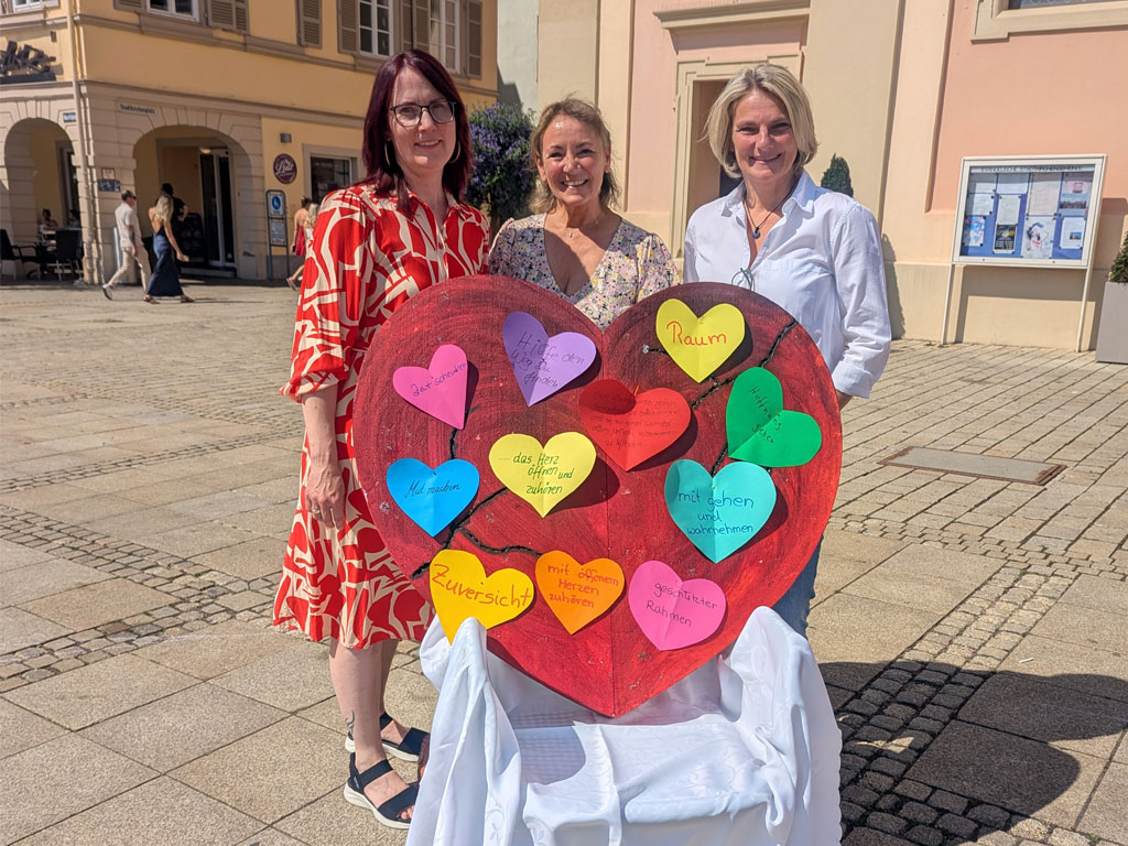 Drei Damen stehen an einem sonnigen Tag auf einem Marktplatz hinter einem weißen Stuhl mit einem Papier-Herz voller herzförmiger Klebezettel.