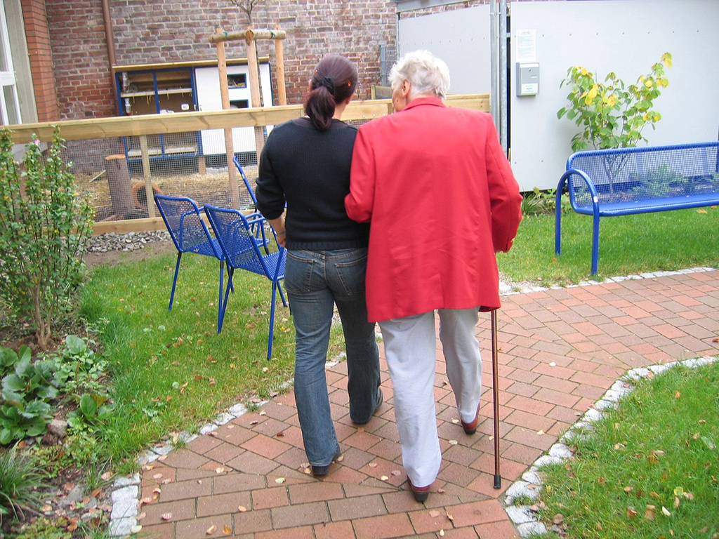 Eine Frau begleitet eine Seniorin mit Stock beim Spazieren gehen